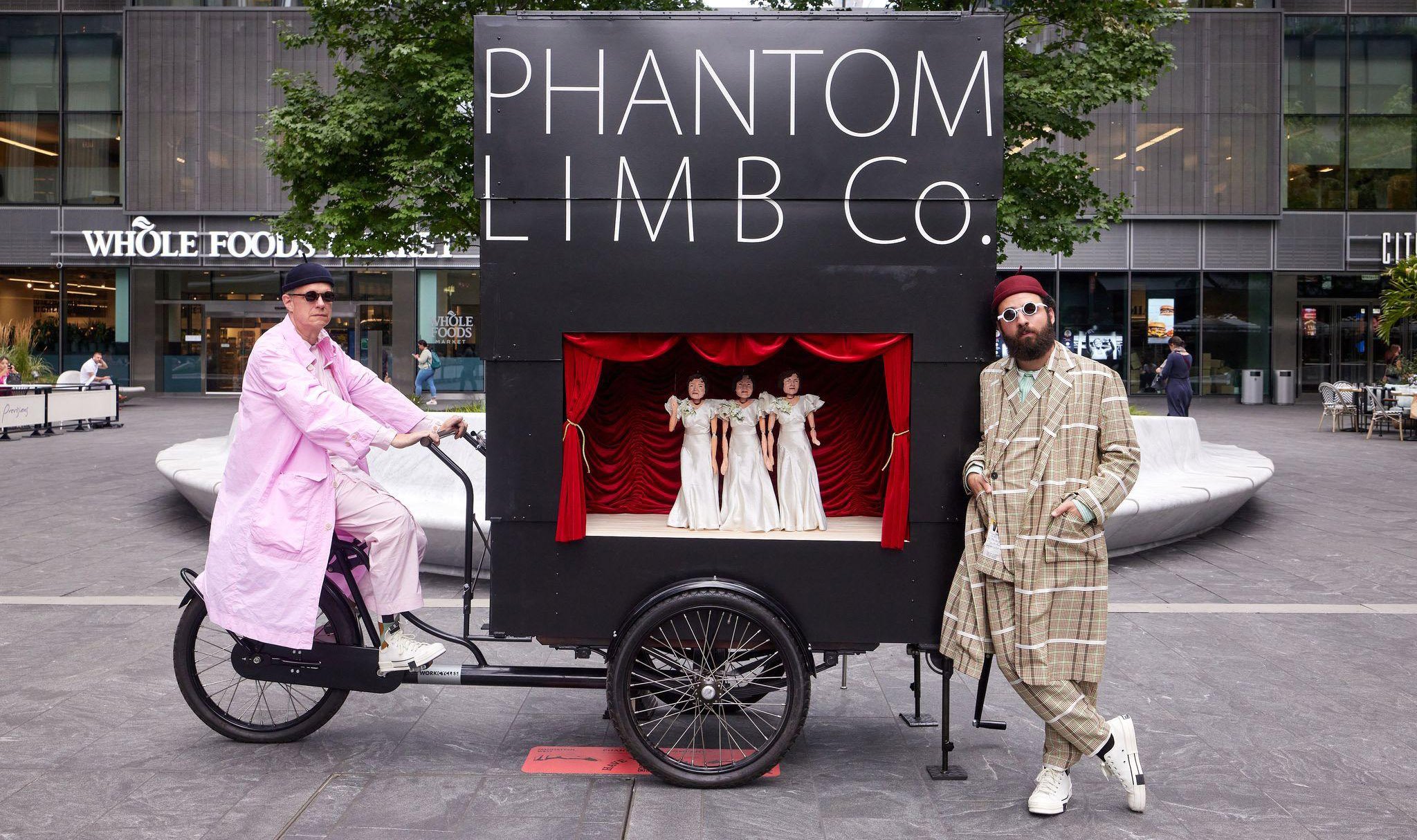 A man posed in a pink trench coat, black beanie, and sunglasses, seated atop cargo bicycle with a large black puppet theatre box on it that says Phantom Limb Co. Another man in a beige plaid suit, red beanie and white sunglasses is leaning against the structure, in the middle of an urban plaza surrounded by people, trees, and buildings.