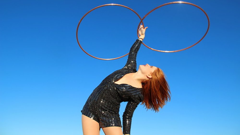 A red-headed white woman leaning the top half of her body over to the right, one arm extended straight to the sky, spinning two hoops, while wearing a sparkly black leotard against a clear blue sky