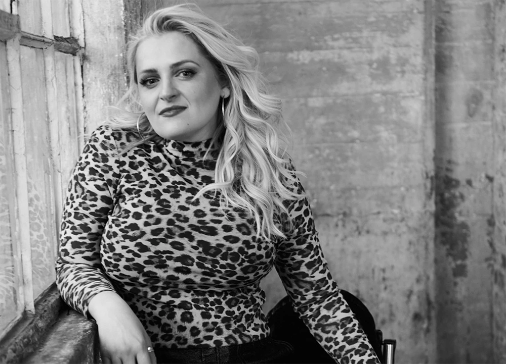 A black & white headshot of Ali Stroker in her wheelchair leaning against a window ledge in an industrial building. She is wearing a cheetah print top and hoop earrings.
