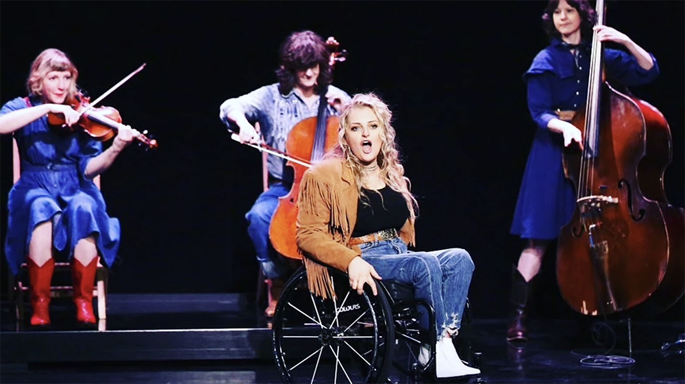 Ali Stroker singing on stage in her wheelchair and western-style costuming, with three string musicians accompanying her in the background.