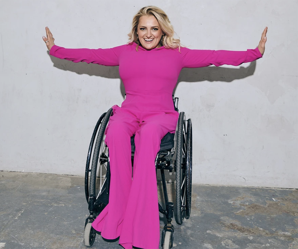 A headshot of Ali Stroker in her wheelchair in a bright pink jumpsuit with arms outstretched to either side.