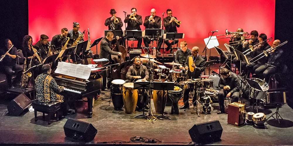 Arturo O'Farrill at the piano, on stage with the 18-piece Afro Latin Jazz Orchestra, seated in front of a bright red background.