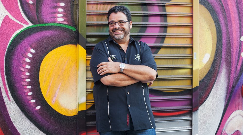 Portrait of Arturo O'Farrill, a brown-skinned man with dark hair, glasses, and facial hair, arms crossed, standing against a brightly colored graffiti wall, wearing a blue short-sleeved collared button down shirt.