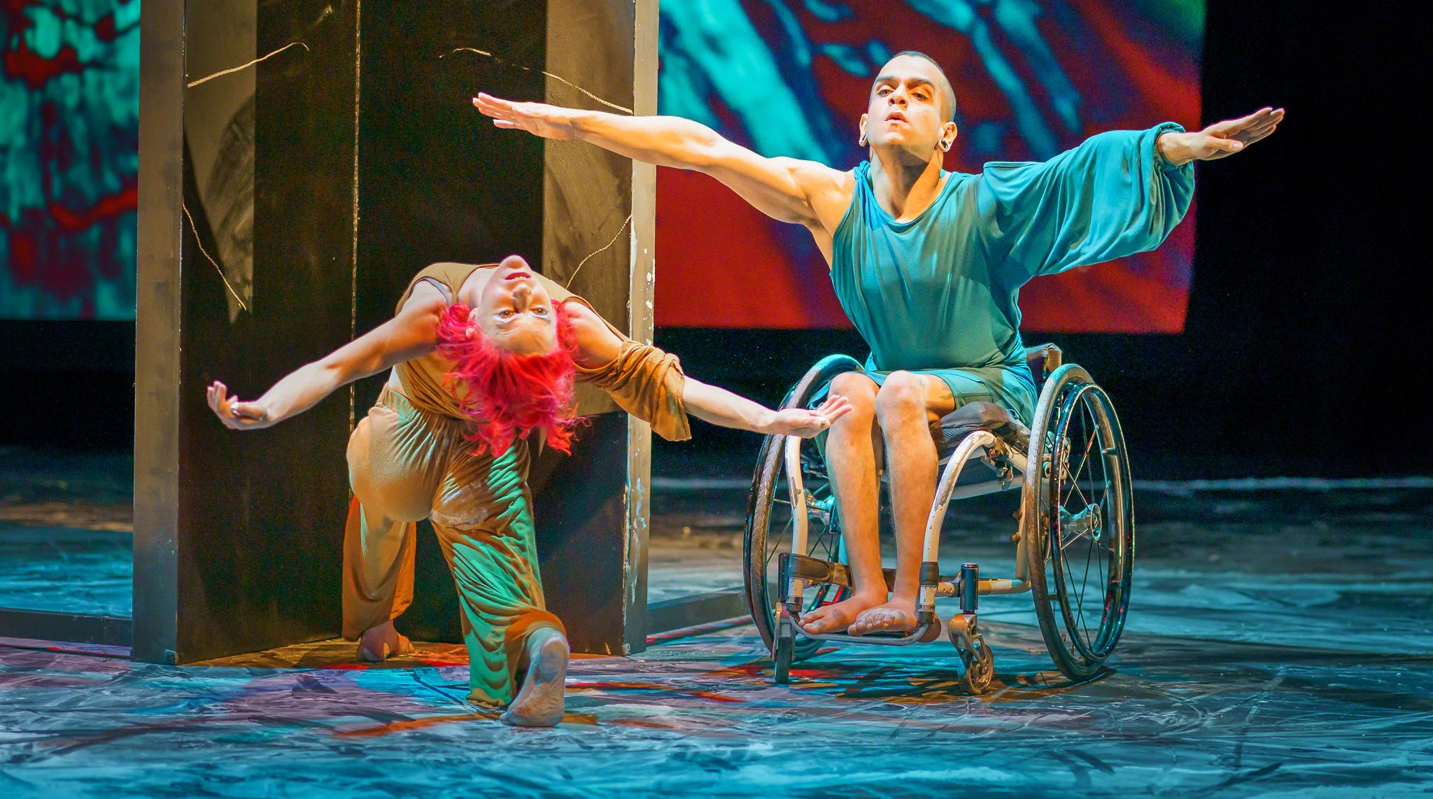 Two dancers on a vibrantly lit stage, one woman on a knee, back toward the audience, in an exxagerated backbend, with bright fuschia hair. The male dancer is in a wheelchair with a focused gaze, arms extended and fingers pointing out.