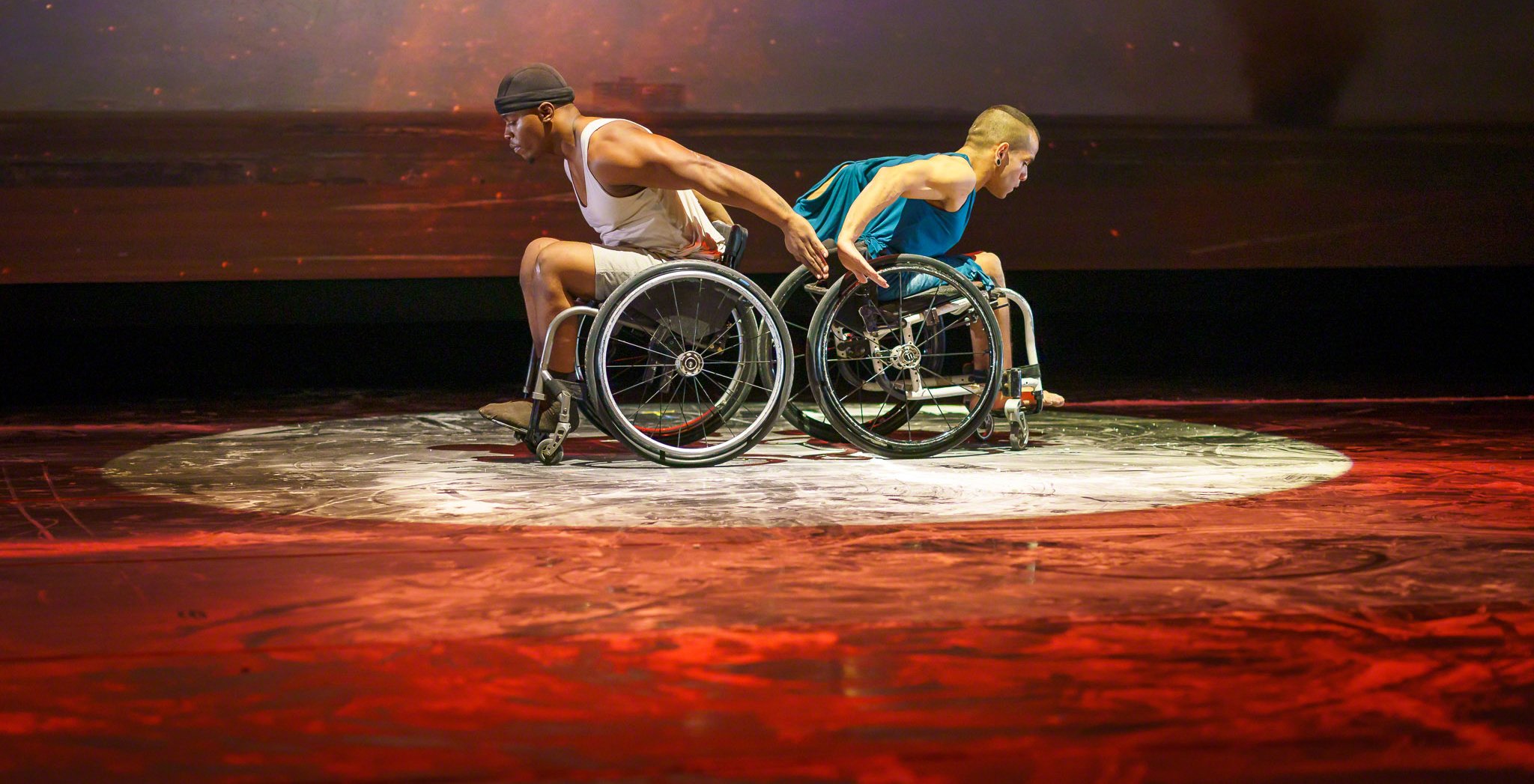 Two men dancers in wheelchairs on stage in a pool of light with their backs together.
