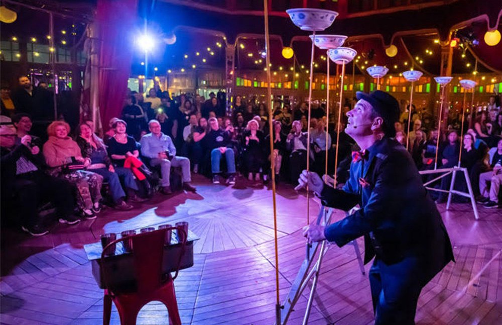 A white man in a navy suit and round top hat balancing bowls on tall sticks while a crowd watches under a red circus tent