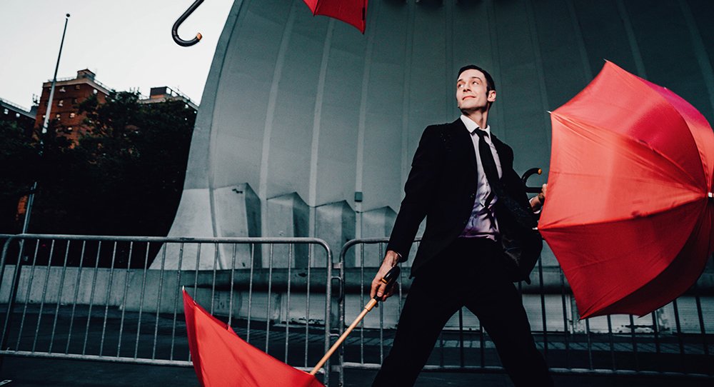 A white man in a black suit juggling opened red umbrellas outside in front of a grey stone amphitheatre