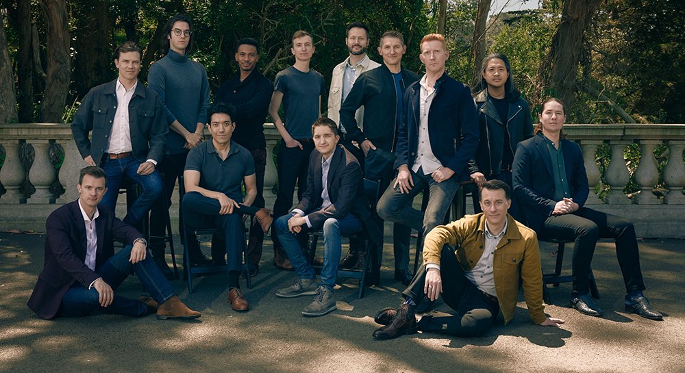 12 male members of Chanticleer in casual sport coats, button down shirts, and khaki pants posed outside against trees on a patio with a pretty garden wall behind them