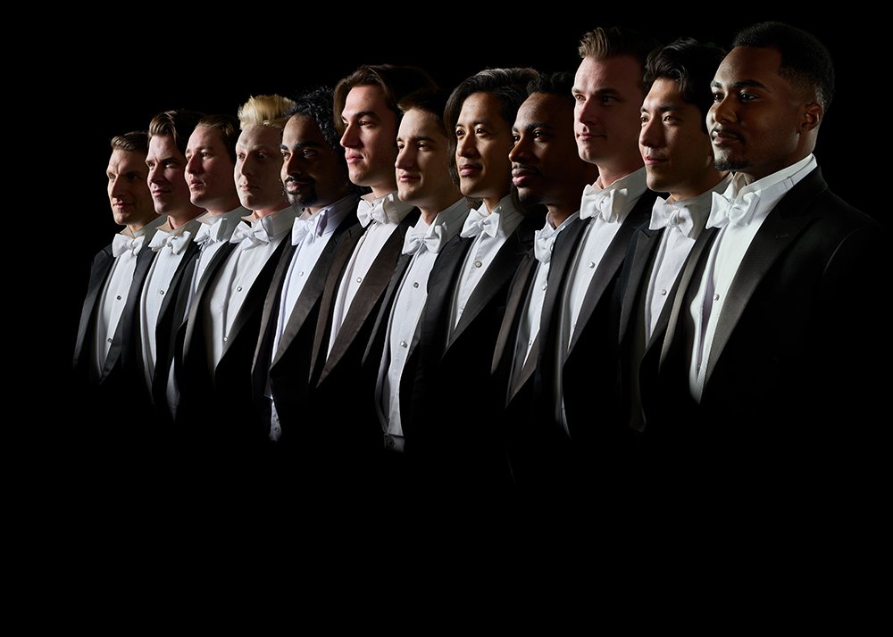 Group profile headshot of 12 male members of Chanticleer posed in a line in formal tuxedos against a black background
