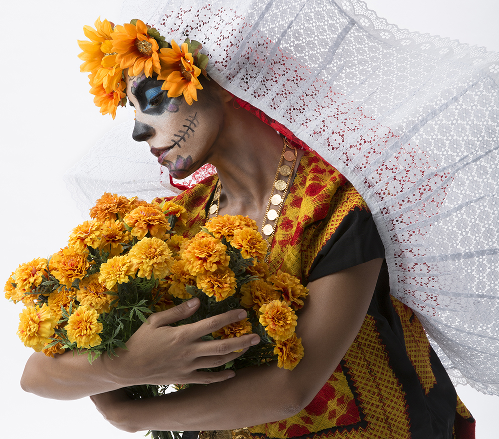 1. Closeup side profile of a woman with sugar skull face makeup, a marigold-lined white-lace veil, holding a bouquet of marigolds, which she is looking down upon, solemnly.