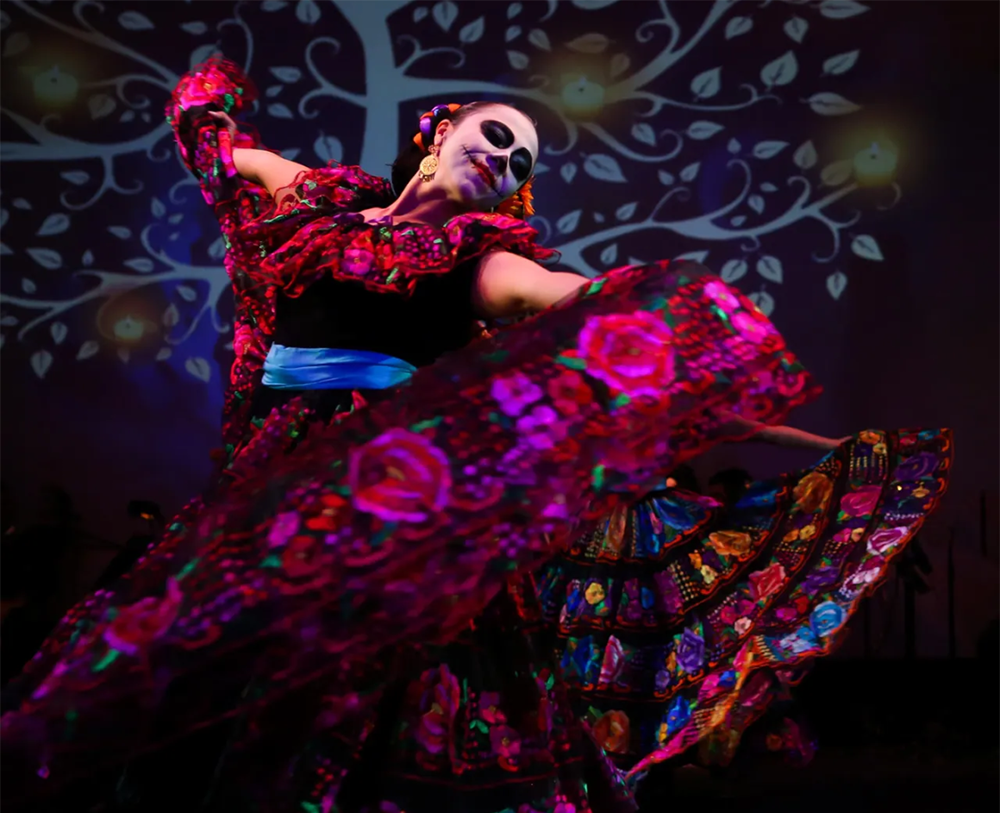 3. A sugar skull dancer, swinging her magenta skirt on a stage in front of a folk art tree projection.