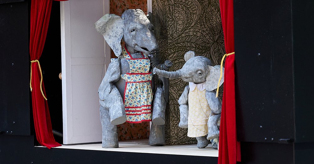 Close-up of a small puppet theatre with two elephant marionettes in dresses, framed by red curtains.