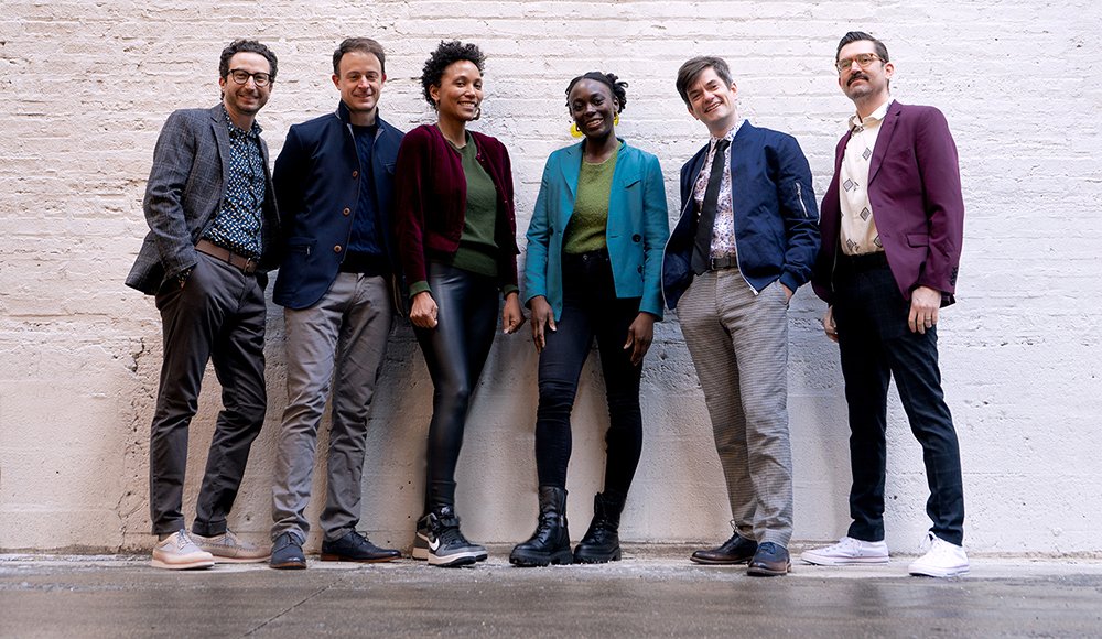 Four white male members of Third Coast Percussion wearing different colored sport coats, standing against a white brick wall with two Black women members of Flutronix