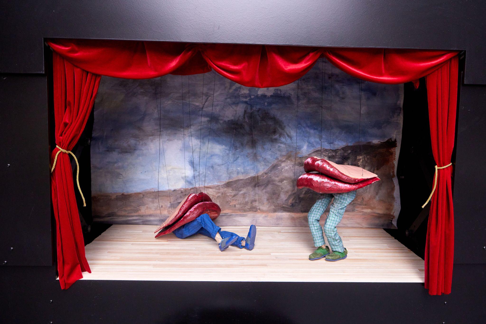 Close-up of a small black puppet theater, with red curtains, and two large puppet lips frowning, with legs