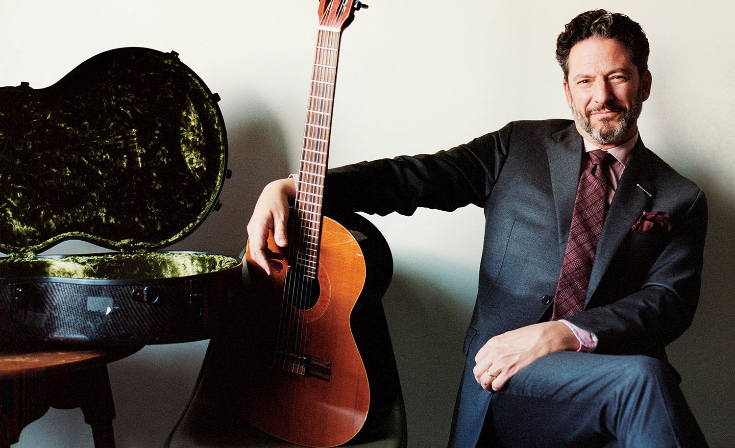 John Pizzarelli seated in a blue suit, right arm outstretched around his guitar, positioned upright, posed next to a rugged, open guitar case.