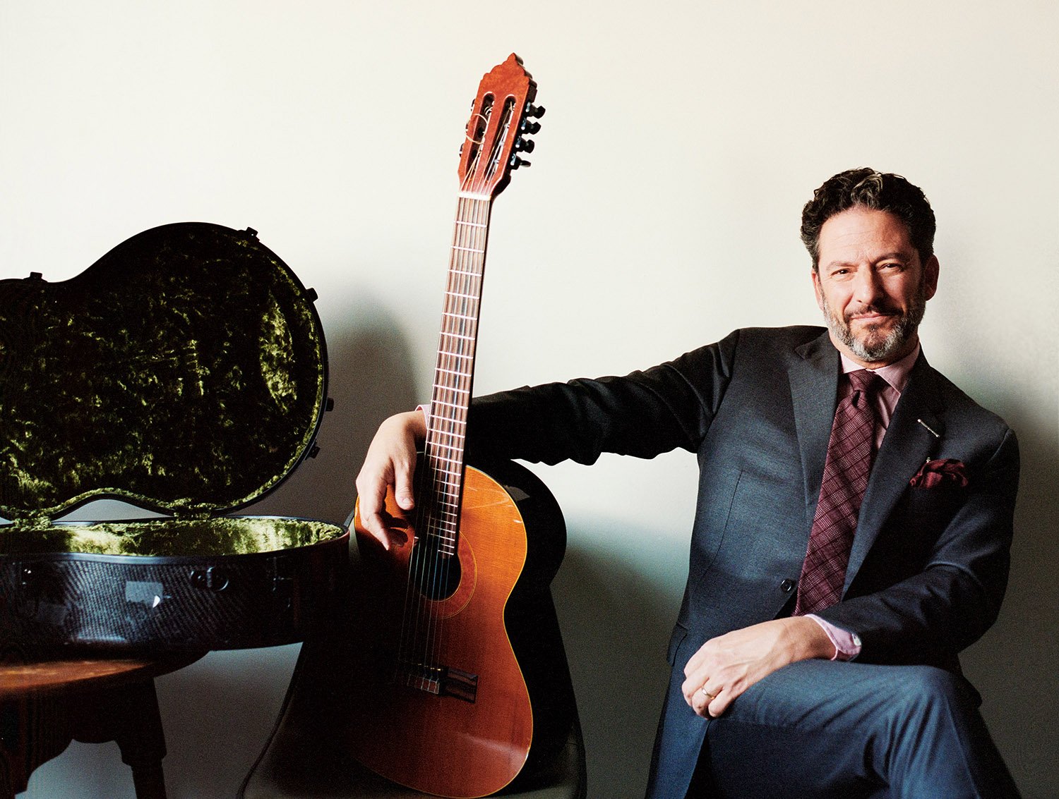John Pizzarelli seated in a blue suit, right arm outstretched around his guitar, positioned upright, posed next to a rugged, open guitar case.