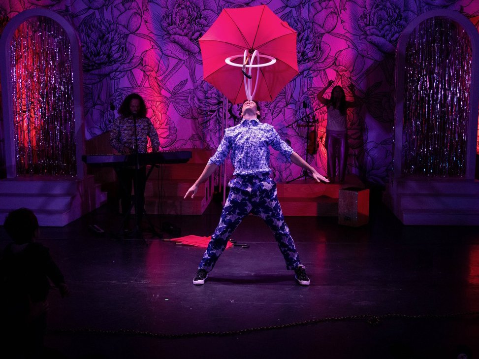 A white man balancing  round tubes and an umbrella in his mouth on stage in front of a sparkly backdrop