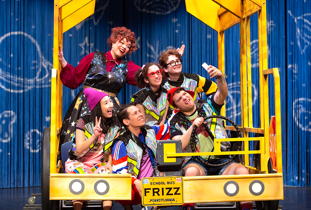 Cast of six actors, five adults costumed as school children, and one dressed as the quirky teacher, Miss Frizzle, posing for a group selfie inside of the school bus theatrical set, with a solar system backdrop.