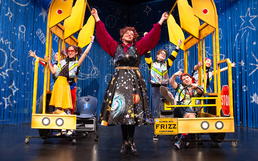 Miss Frizzle standing in the center with arms raised, hands in fists, with “students” on either side, posing victoriously.