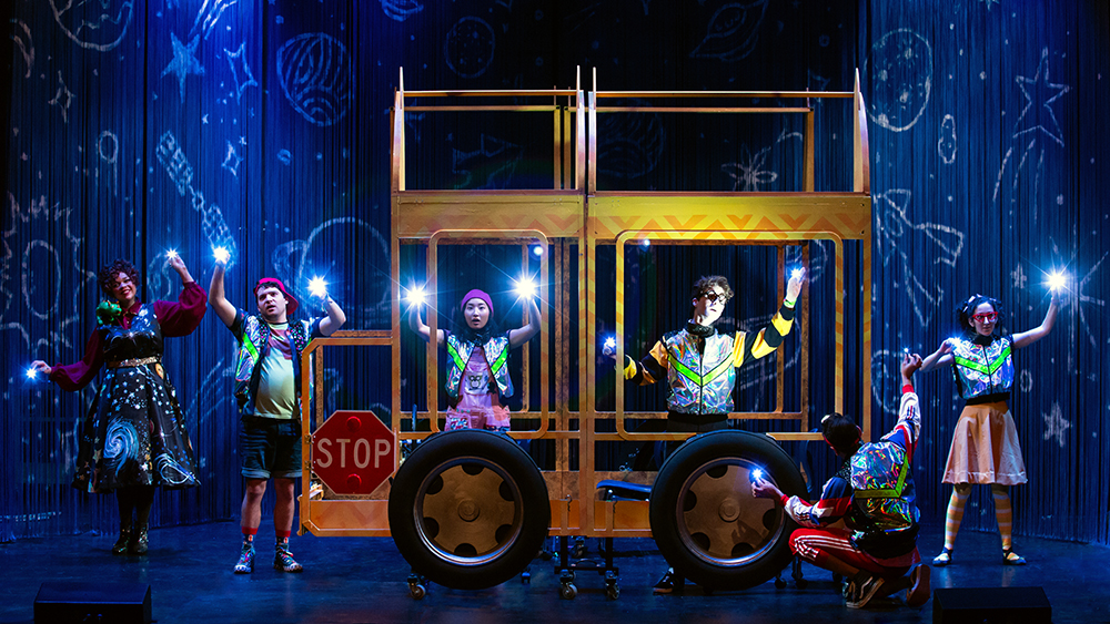 The cast of the Magic School Bus, posed on a dimly-lit stage, pointing flashlights toward the audience.