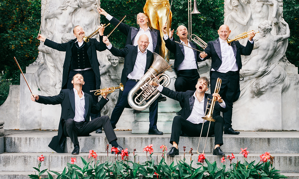 Seven men in concert attire holding a variety of brass instruments, some also with strings bows in hand, posing in a garden in front of a gold statue of Johann Strauss.