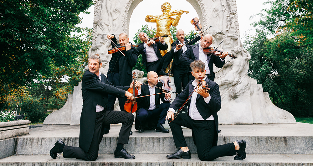 Seven men in concert attire with a variety of silly facial expressions holding violins with incorrect form in garden in front of a gold statue of Johann Strauss.