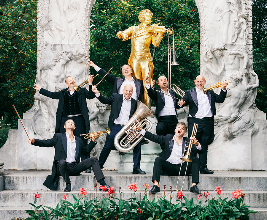 Seven men in concert attire with silly smiles holding a variety of brass instruments, some also with strings bows in hand, posing in a garden in front of a gold statue of Johann Strauss.