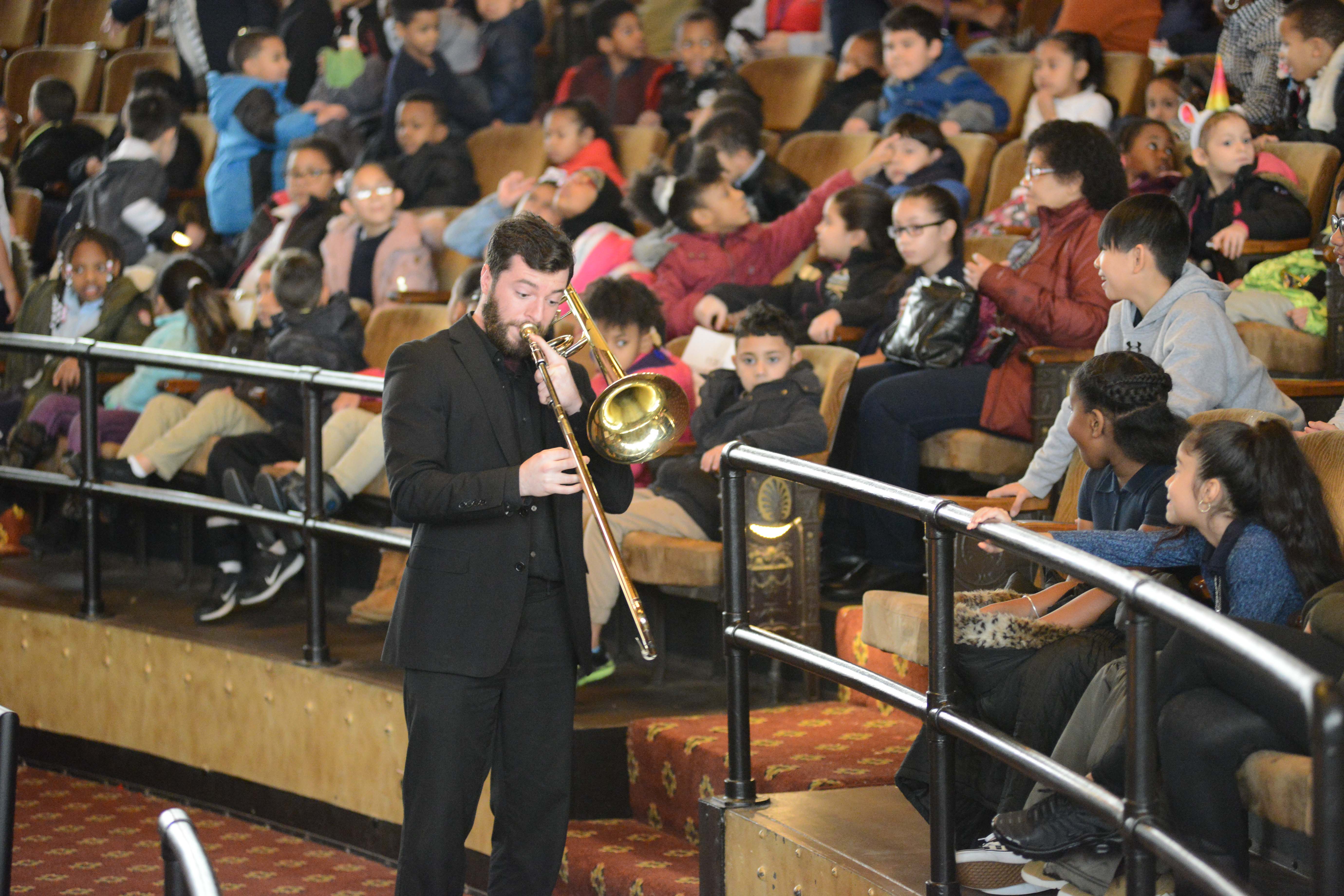 A musician playing a trombone walking through an auditorium filled with children