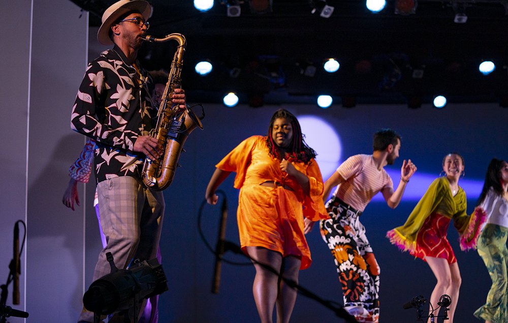 A Black man in the foreground playing saxophone with a straw top hat, black and white floral blouse, and checked pants, with four diverse tap dancers in bright outfits dancing and smiling to his music.