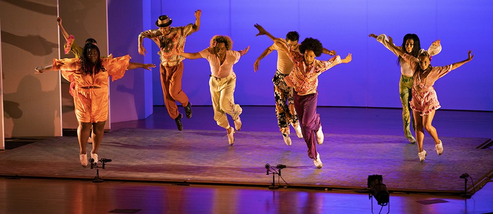 Seven tap dancers are mid jump on stage, arms outstretched, with a purple light as background.