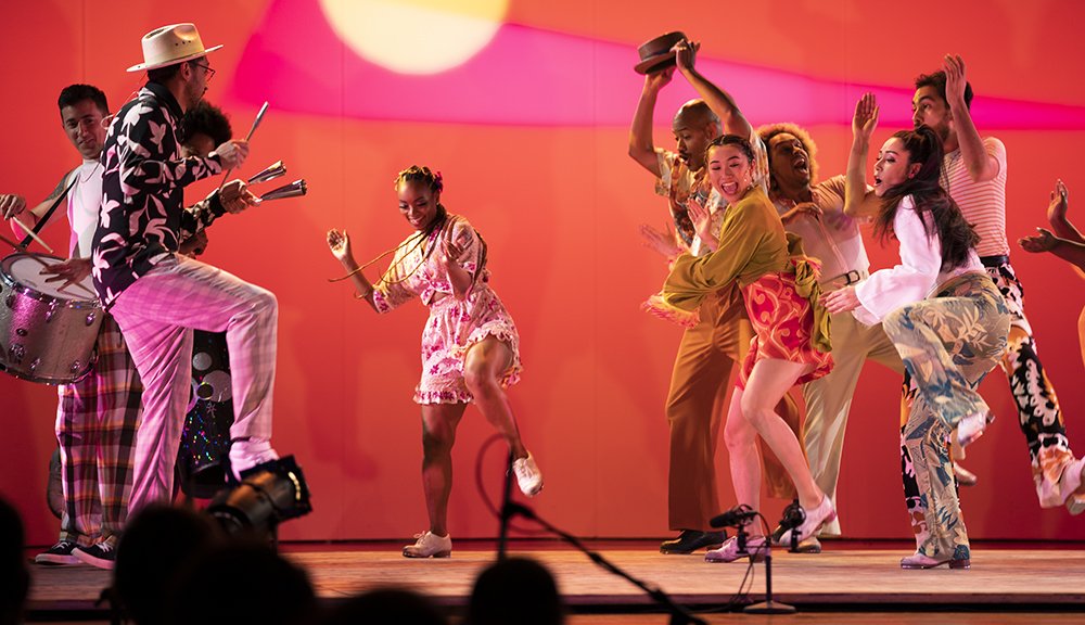 An ensemble of seven black and brown tap dancers, in a variety of poses and outfits, dancing on stage behind a vibrant, geometric sunset projected behind them.