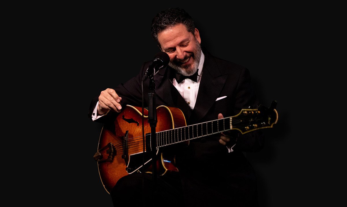 John Pizzarelli in a tuxedo with bowtie, performing seated in front of a mic, mid-guitar strum