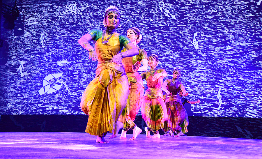 1. Four Bharatanatyam dancers in colorful costumes in a line, dancing in front of a blue and white projected indigenous Warli painting.