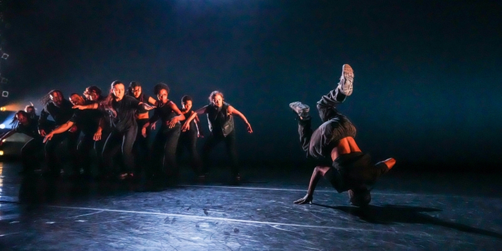 A breakdancer on stage, spinning on his head, while a cluster of dancers stage right look on.