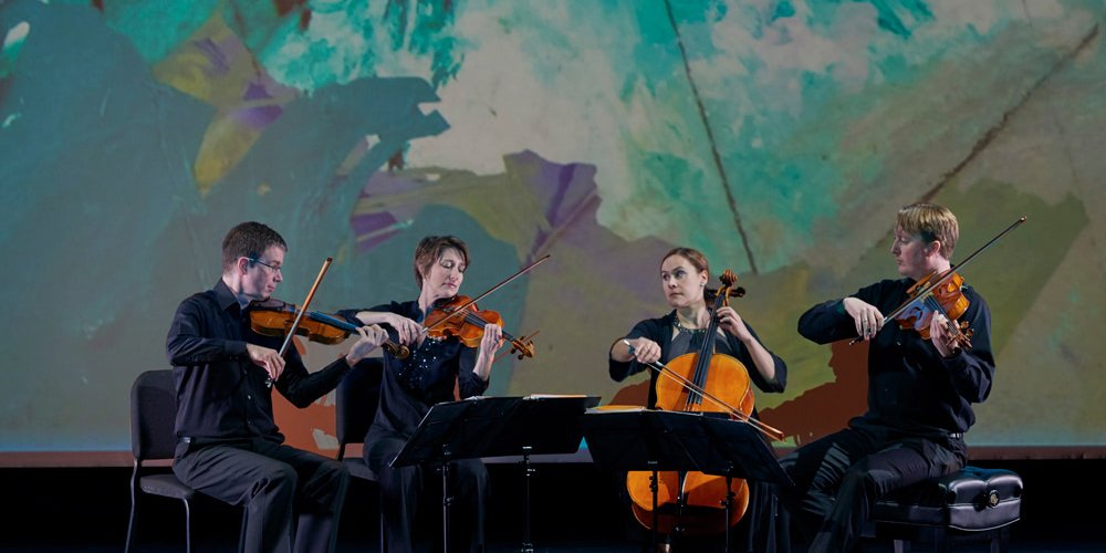 A chamber quartet performing on stage with a painting of ocean waves projected behind them.