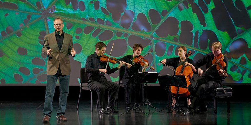 A chamber quartet performing on stage with a man in a suit speaking to the audience, and a vibrant, close-up photo of a green leaf with lots of holes, projected behind them.