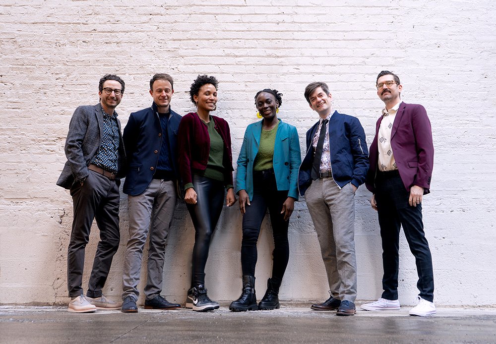 Four white male members of Third Coast Percussion wearing different colored sport coats, standing against a white brick wall with two black women members of Flutronix