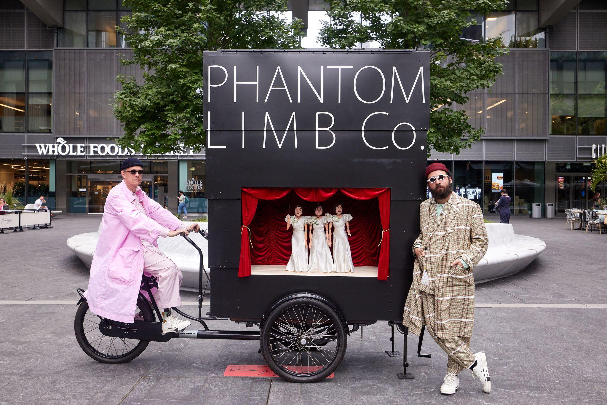 A man posed in a pink trench coat, black beanie, and sunglasses, seated atop cargo bicycle with a large black puppet theatre box on it that says Phantom Limb Co. Another man in a beige plaid suit, red beanie and white sunglasses is leaning against the structure, in the middle of an urban plaza surrounded by people, trees, and buildings