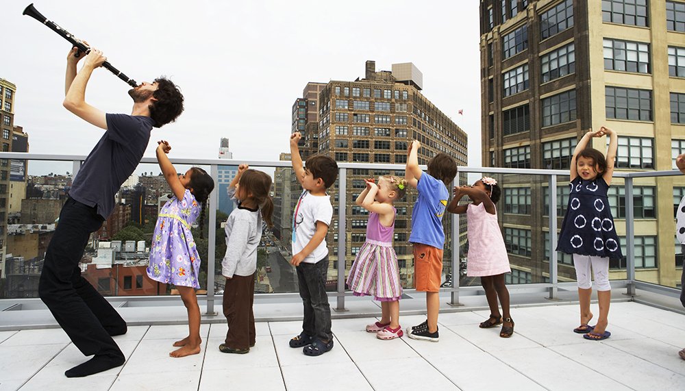 Musician Oran Etkin is on an urban rooftop dress in dark colors playing his clarinet pointed to the sky, with a line of kids behind him, with their arms raised, in a "pied piper" scene