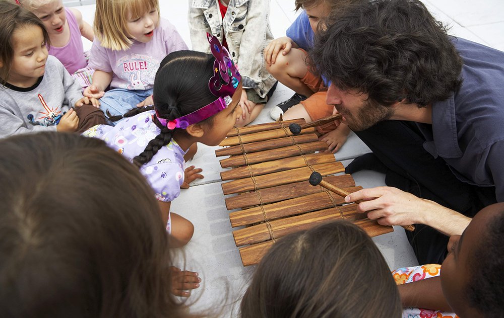 Musician Oran Etkin sitting on the floor playing a xylophone surrounded by children looking on in wonder.