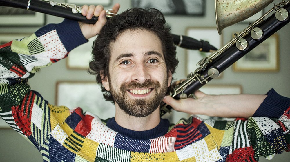 Close-up shot of musician Oran Etkin wearing a brightly colored and patterned sweater and holding a clarinet and saxophone over his head.