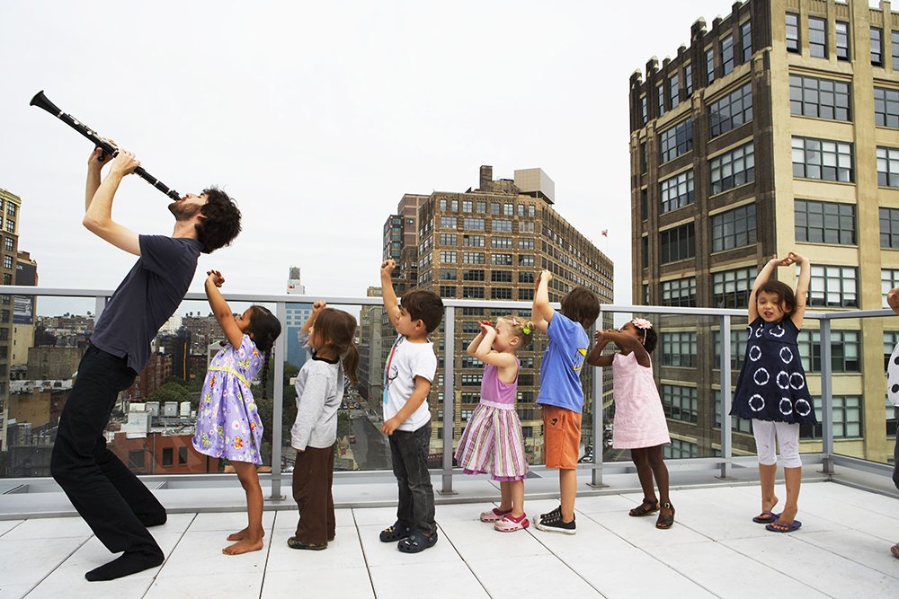 A white male musician playing his clarinet to the sky on a city rooftop, followed by children in a line behind him