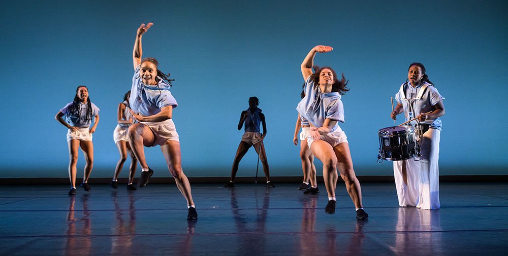 Five Black women dancers in white shorts, black sneakers, and light blue sleeveless, hooded, crop tops in an active, mid-jump dance move on stage, accompanied by a percussionist with drum around her neck.