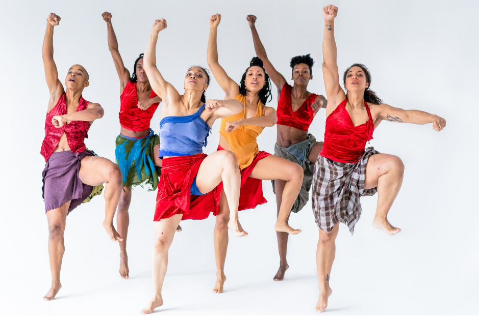 6 strong dancers with one leg and one fist raised in an empowered posture, wearing red lux halter tops, and skirts made from a variety of different fabrics.