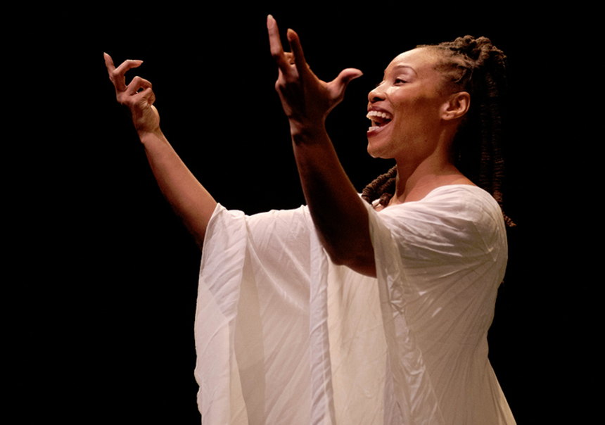 Side profile of a Black woman in a flowy-sleeved white dress, both arms up and open, with palms and fingers stretched to the sky, a joyful expression on her face