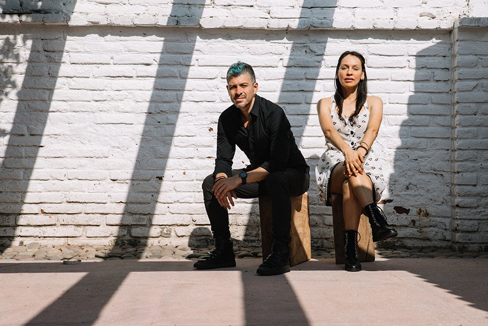 Rodrigo and Gabriela sitting on a bench outside in front of a white brick wall with shadows creating stripes over the scene