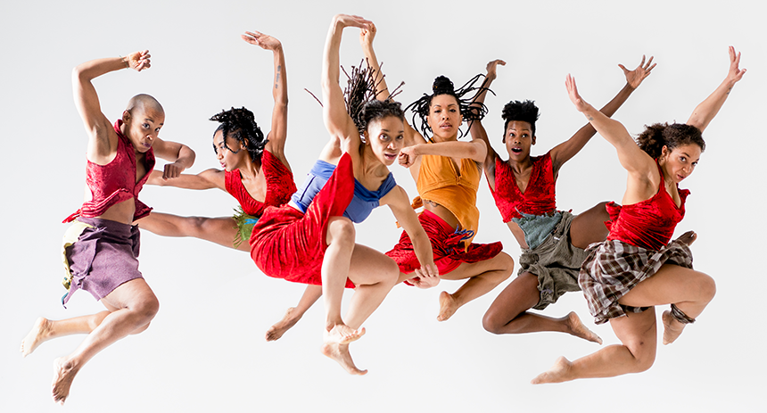 6 strong dancers jumping in mid-air, arms stretch to the sky, wearing red lux halter tops, and skirts made from a variety of different fabrics.